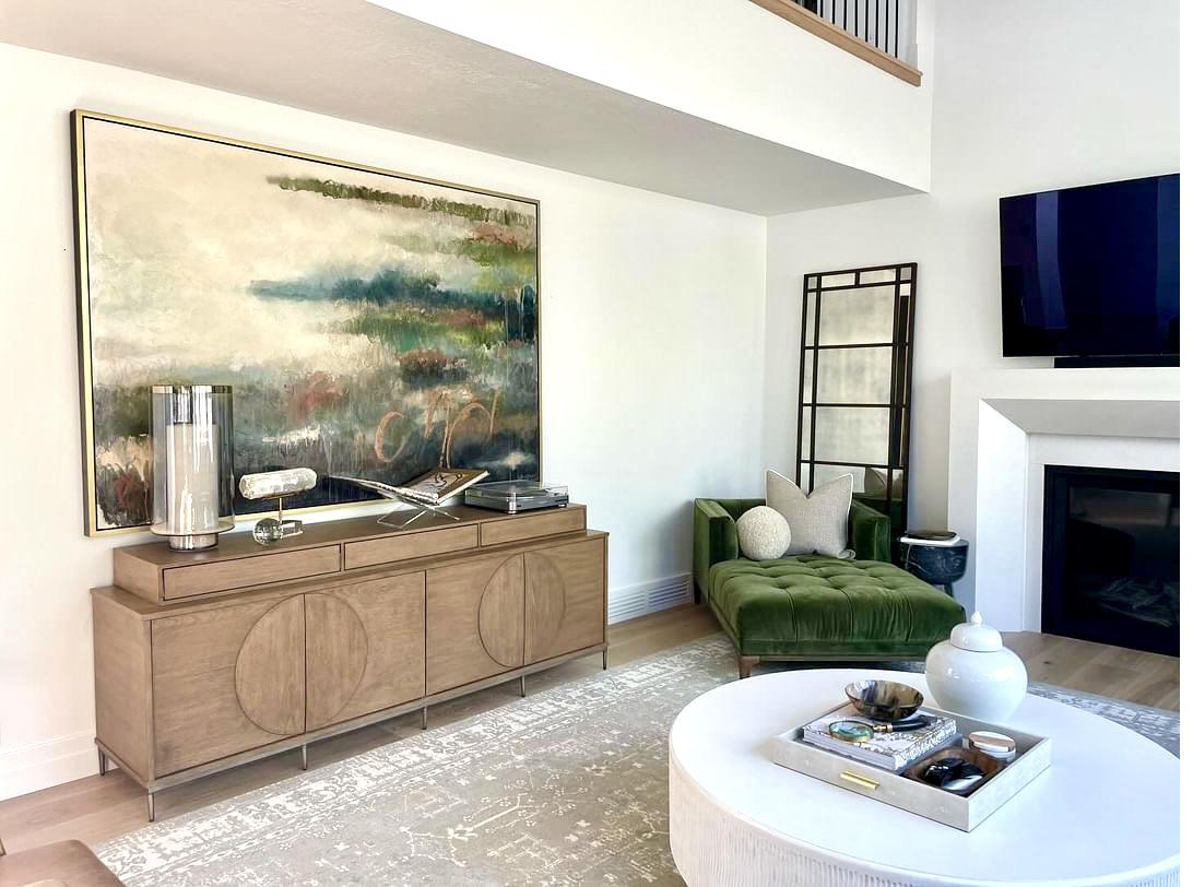Large abstract landscape painting in greens teals and earth tones displayed above wooden credenza in bright modern living room with green velvet chaise lounge
