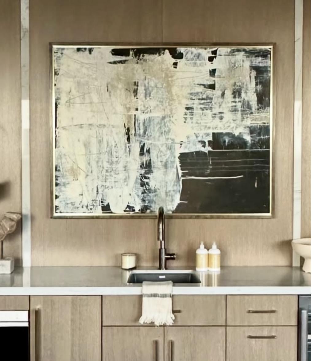 Large abstract painting in black charcoal and white with gestural brushstrokes in gold frame displayed above modern kitchen sink with oak cabinetry
