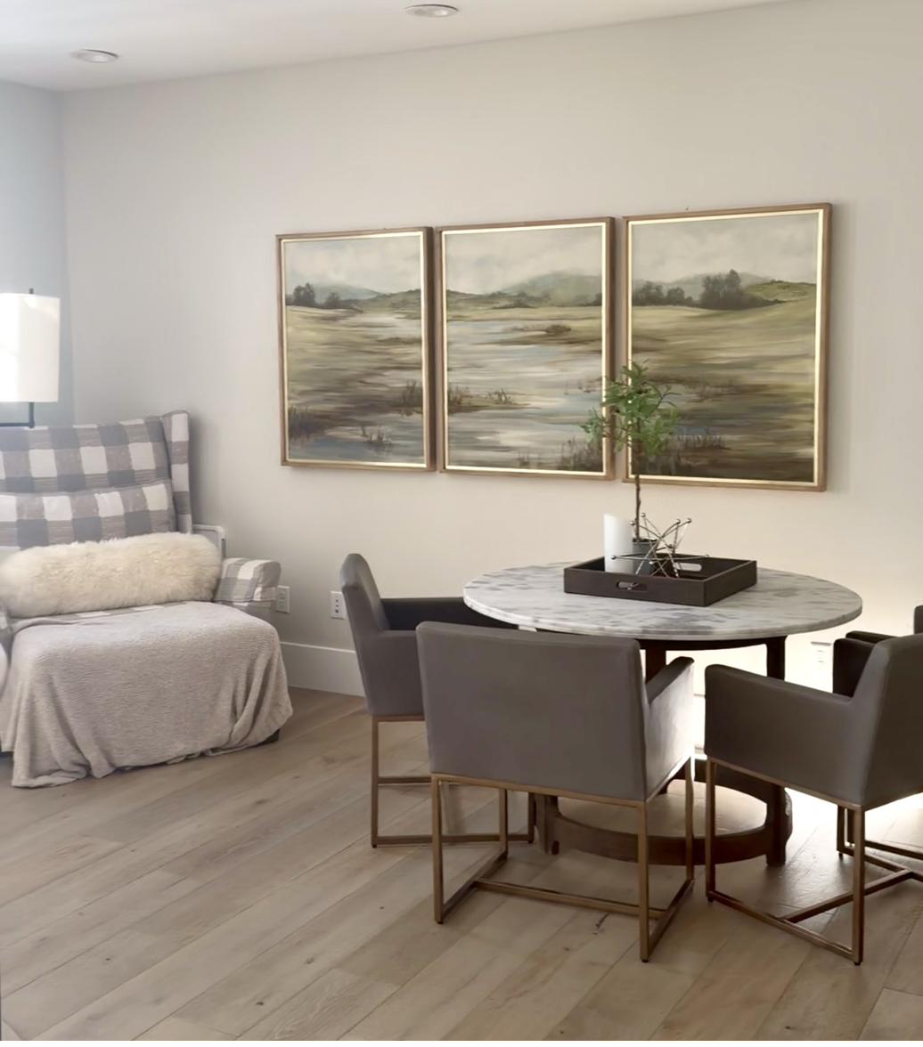 Three-panel landscape triptych painting in soft greens and muted tones depicting rolling hills and water in gold frames displayed above round marble dining table