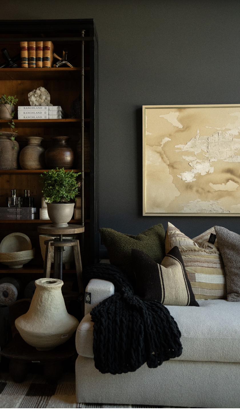 Warm-toned textured abstract painting in cream and sienna with dimensional plaster technique on gold frame displayed above sofa in moody living room with vintage bookcase