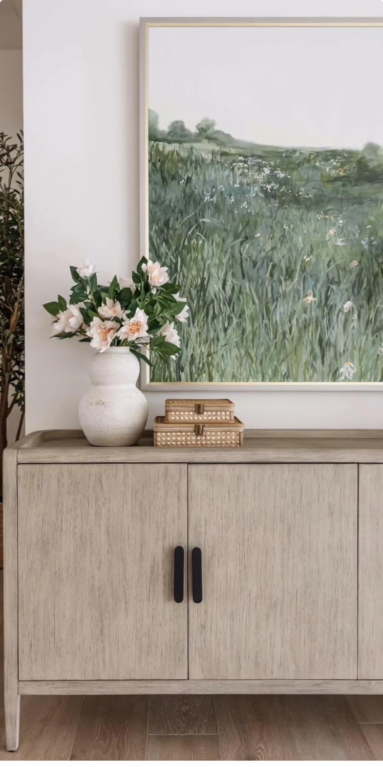 Large painting of lush green wildflower meadow in gold frame displayed above light oak console table with ceramic vase and pink flowers in bright entryway