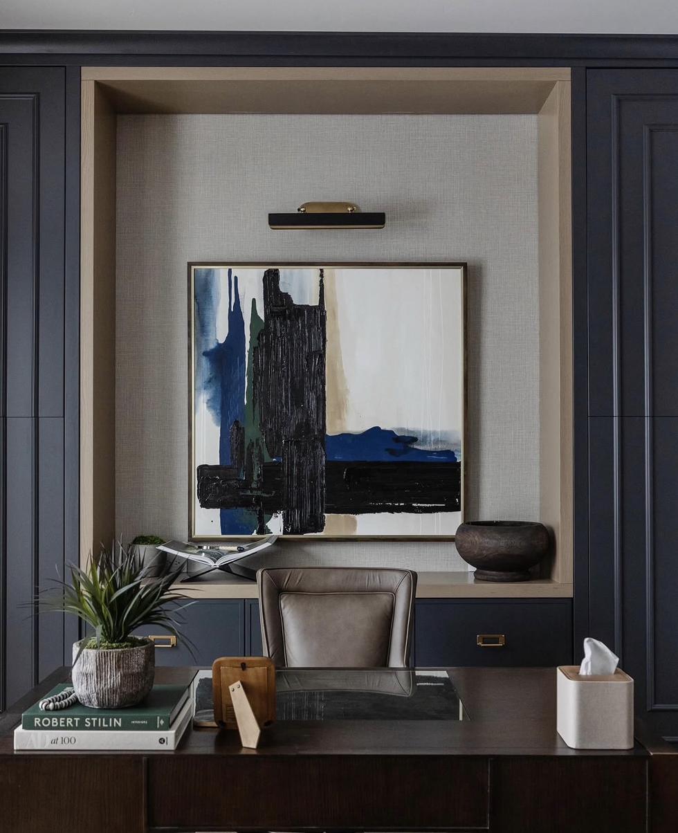 Bold abstract painting with black and blue brushstrokes displayed in sophisticated home office with navy blue paneled walls and brass picture light