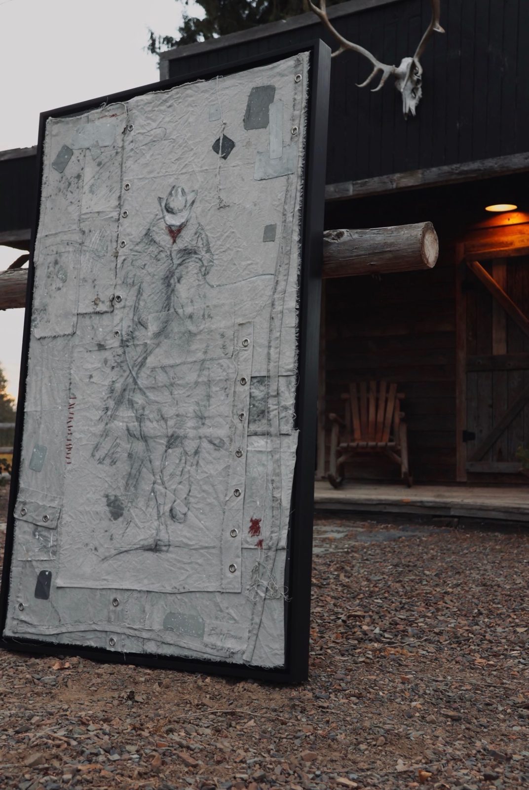 Large mixed media piece of a sketched cowboy on a patchwork of stitched and grommeted canvas, leaning against a rustic log cabin