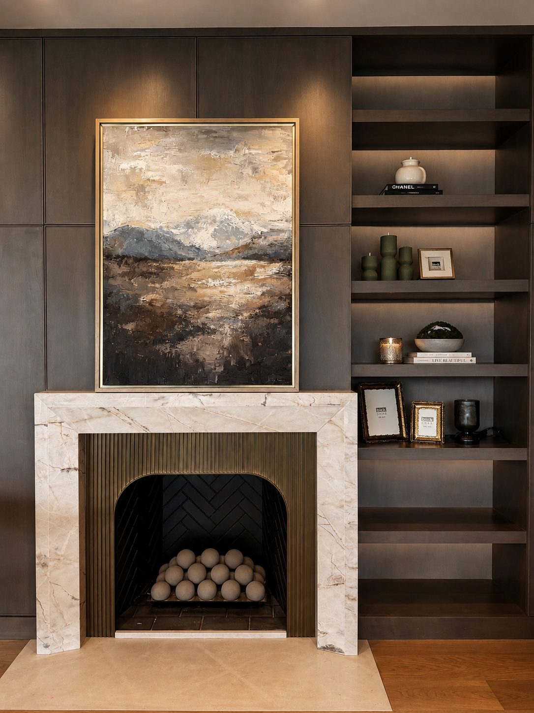 Moody mountain landscape painting in earth tones with snow-capped peaks, in a gold frame above a marble fireplace with dark built-in shelving